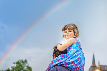 Little preschool girl sitting on shoulder of father. Happy toddler child and man observing rainbow on sky after summer rain. Happy family, bonding, love. Summertime. Dad and daughter. Father's day.