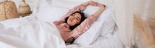 Young Asian Woman In Silk Pajamas Waking Up With Outstretched Hands And Closed Eyes In Bedroom, Banner