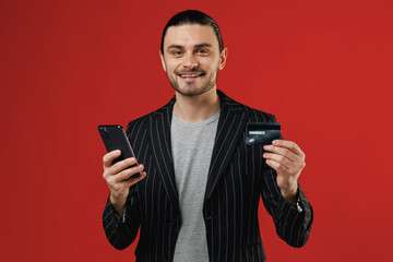 Wall Mural - Young businessman latin man in black striped jacket grey shirt using mobile cell phone hold credit bank card doing online shopping order delivery booking tour look camera isolated on red background