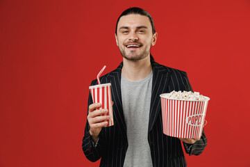 Wall Mural - Young cheerful friendly happy smiling fun successful businessman latin man 20s wear black striped jacket grey shirt holding takeaway popcorn bucket soda cola isolated on red color background studio