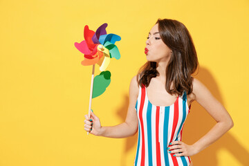 Happy young sexy woman slim body wear striped red blue one-piece swimsuit hold blow on toy windmill isolated on vivid yellow color wall background studio. Summer hotel pool sea rest sun tan concept