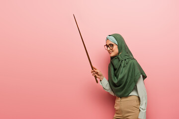 Wall Mural - smiling muslim teacher looking away while holding pointing stick on pink background