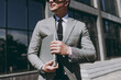 © ViDi Studio - Close up cropped photo portrait shot successful young employee business man 20s in grey suit stand near office glass wall building outdoors in downtown city hold closed laptop pc computer look aside.