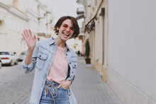 Young Smiling Happy Student Caucasian Woman 20s Wearing Casual Jeans Clothes Eyeglasses Strolling Walk Go Down City Street Outdoors Waving Hand Say Hello People Youth Active Urban Lifestyle Concept