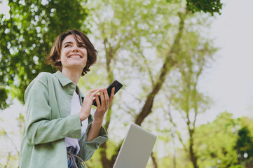 Bottom view young student freelancer woman in green jacket jeans sit on bench in spring park outdoors rest use laptop pc computer talk by mobile cell phone look aside. People urban lifestyle concept.