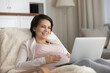 © fizkes - Smiling pregnant woman using laptop, looking at screen, touching caressing belly, sitting in cozy chair at home, happy young future mom watching movie or making video call, shopping online
