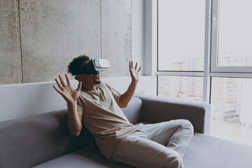 Wall Mural - Side view cool young african american man 20s wearing beige t-shirt sit on grey sofa indoors apartment watch in vr headset pc gadget playing game with outstretched hand rest on quarantine stay home