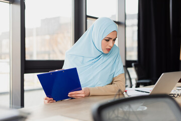 Wall Mural - pretty muslim businesswoman holding clipboard while looking at laptop, blurred foreground