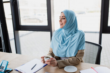 Wall Mural - smiling muslim businesswoman with cup of coffee sitting at workplace against window