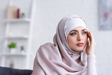 Pensive Muslim Woman In Hijab Holding Hand Near Face While Looking Away At Home