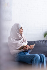 Wall Mural - young muslim woman sitting with book on couch at home on blurred foreground