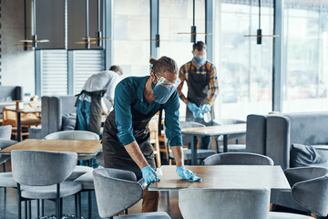 Three young male waiters in protective workwear cleaning tables for clients in restaurant