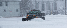 Black Truck With A Snowplow Clearing A Parking Lot Behind Buisnesses