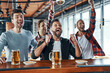 © gstockstudio - Cheering young men in casual clothing drinking beer and watching sport game while sitting in the pub