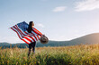 Beautiful young american brunette woman in hat standing on grass of background mountains in wind independent and young, holds proudly usa flag high in air, demonstrates patriotism and freedom