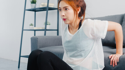 Young Korean lady in sportswear exercises doing working out doing tricep dips leaning on couch in living room at home. Social distance, Isolation during the virus. Exercises for the lower body.