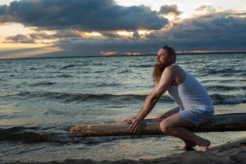 Wall Mural - Funny bald man with red beard posing on the beach at sunset. A humorous male parody of a glamorous girl.