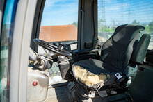 Old Empty Shabby Driver's Seat In The Cab Of A Tractor, Crane Or Other Agricultural Or Construction Vehicle With A Steering Wheel And Large Windows On A Sunny Summer Day At A Farm Or Construction Site