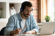 © fizkes - Happy focused young man in eyeglasses wearing wireless headphones, watching educational lecture on computer, studying on online courses distantly, improving professional knowledge, writing notes.