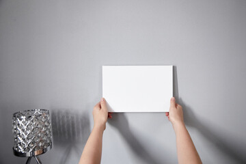 White canvas in female hands with gray wall background. Woman hanging blank picture mockup on wall