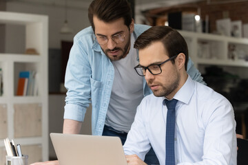 Canvas Print - Concentrated two colleagues looking at laptop screen, discussing online project, preparing electronic documents, developing strategy. Skilled male employee helping coworker with corporate software app