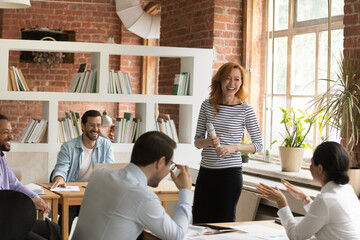 Wall Mural - Happy young friendly red-haired female speaker with microphone in hands leading negotiations meeting or giving educational lecture seminar to relaxed positive motivated multiracial business people.