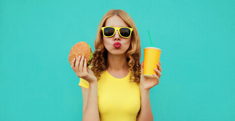 Wall Mural - Portrait close up of young woman with fast food, burger and cup of juice wearing a yellow t-shirt, sunglasses on a blue background