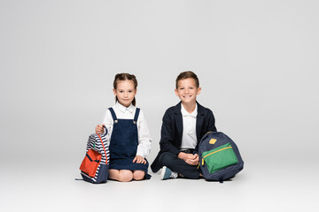 Wall Mural - cheerful schoolkids in uniform sitting with backpacks on grey.