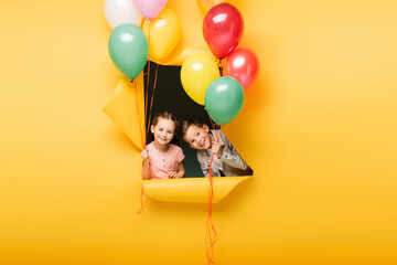 cheerful kids holding colorful balloons through hole on yellow background.