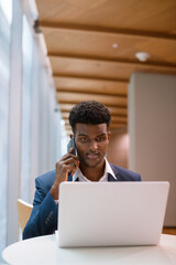 Portrait of African businessman using laptop computer and talking on phone in coffee shop