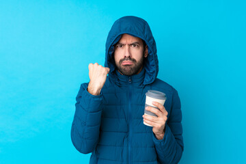 Man wearing winter jacket and holding a takeaway coffee over isolated blue background with angry gesture