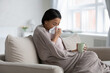 Frustrated sick mixed raced woman wrapped in warm blanket catching cold, suffering from flu illness or infection symptoms, drinking mug of hot herbal tea, blowing nose, taking treatment at home