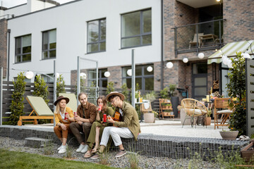 Wall Mural - Group of young friends spending summertime together, having fun sitting together with drinks and guitar on a porch of the country house, wide view on a beautiful cottages