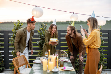 Young adult friends celebrating birthday, having fun blowing candles on a cake at backyard of a country house outdoors