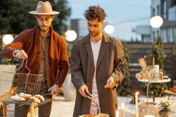 Wall Mural - Two male friends serving grilled sausages on a dining table outdoors. Fun summer time in a country house, male friendship