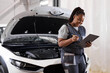 © Roman - black woman mechanic engineer holding checklist paper and taking notes on clipboard, african woman is standing next to car engine hood, wearing uniform overalls. side view portrait