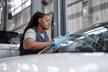 Wall Mural - Afro woman polishing cleaning car with microfiber cloth, car detailing or valeting concept. Side view on black lady in overalls at work. copy space