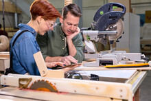 Customer And Joiner Have Talk In Workshop, Discussing, Sharing Ideas What To Make From Wood, Handmate Furniture. Two Caucasian Man And Woman Indoors, Sawing Machine In The Background