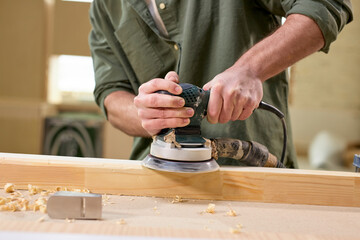 Cropped male carpenter carefully smoothing wooden material with electrical sander, concentrated on work, enjoy woodworking, side view portrait of man making furniture. close-up photo