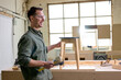 Side view portrait of happy carpenter man in safety goggles using holding hammer in hands during work, having handmade stool chair on workbench, smile. copy space.
