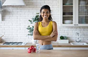 Portrait of African American beautiful young fitness trainer woman with crossed arms, standing at home kitchen, looking at the camera, healthy lifestyle, fitness at home online