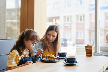 Wall Mural - Warm-toned portrait of caring mother and daughter with down syndrome enjoying cakes in cafe, copy space