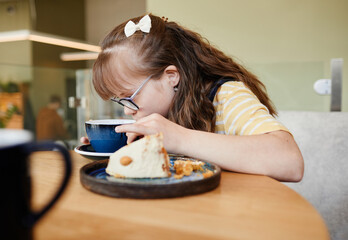 Wall Mural - Candid side view portrait of cute girl with down syndrome enjoying desserts in cafe, copy space