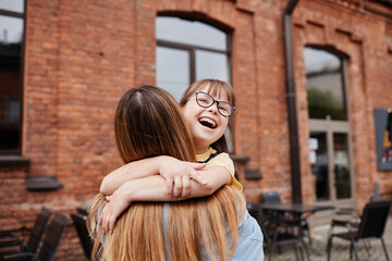 Wall Mural - Candid portrait of carefree girl with down syndrome embracing mother outdoors in city, copy space