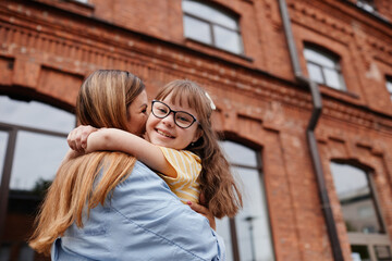 Wall Mural - Candid portrait of happy girl with down syndrome embracing mother outdoors in city, copy space