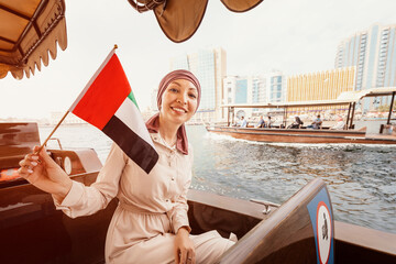 Wall Mural - Happy woman with the flag of the United Arab Emirates sits in a boat during a cruise on the Dubai Creek Canal with views of numerous skyscrapers and attractions