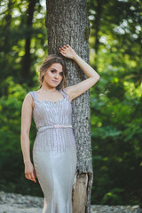 Canvas Print - Attractive Caucasian female wearing an elegant long silver dress leaning on a tall tree in the park