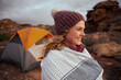 © StratfordProductions - Smiling woman embracing blanket during winter morning wearing cap at mountain trail during camping