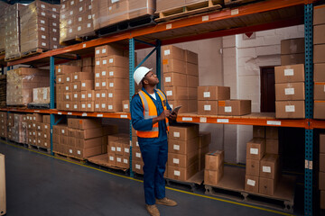 Wall Mural - African male warehouse worker with safety vest and helmet working in large warehouse using digital tablet