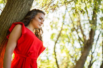 Wall Mural - Portrait of a young caucasian female in a red dress leaning to a tree